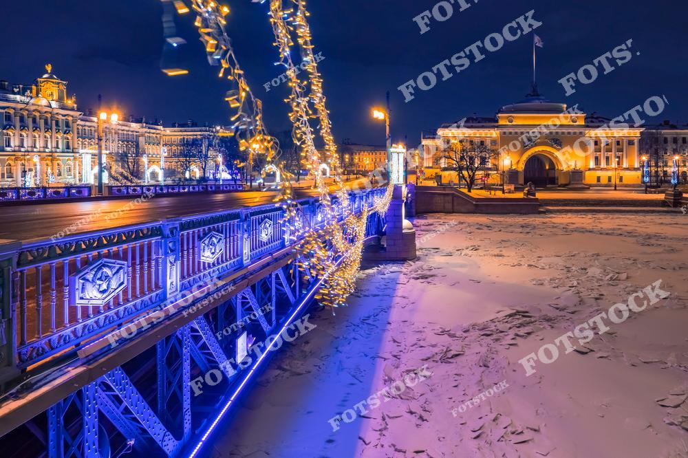 На фотографии изображен Дворцовый мост в зимнюю ночь, украшенный новогодними гирляндами и подсветкой. Ледяные куски на замерзшей Неве подчеркивают зимнюю атмосферу. На заднем плане видны здание Адмиралтейства и Эрмитаж, освещенные теплым светом. Это место является одним из самых красивых и посещаемых в центре Санкт-Петербурга.